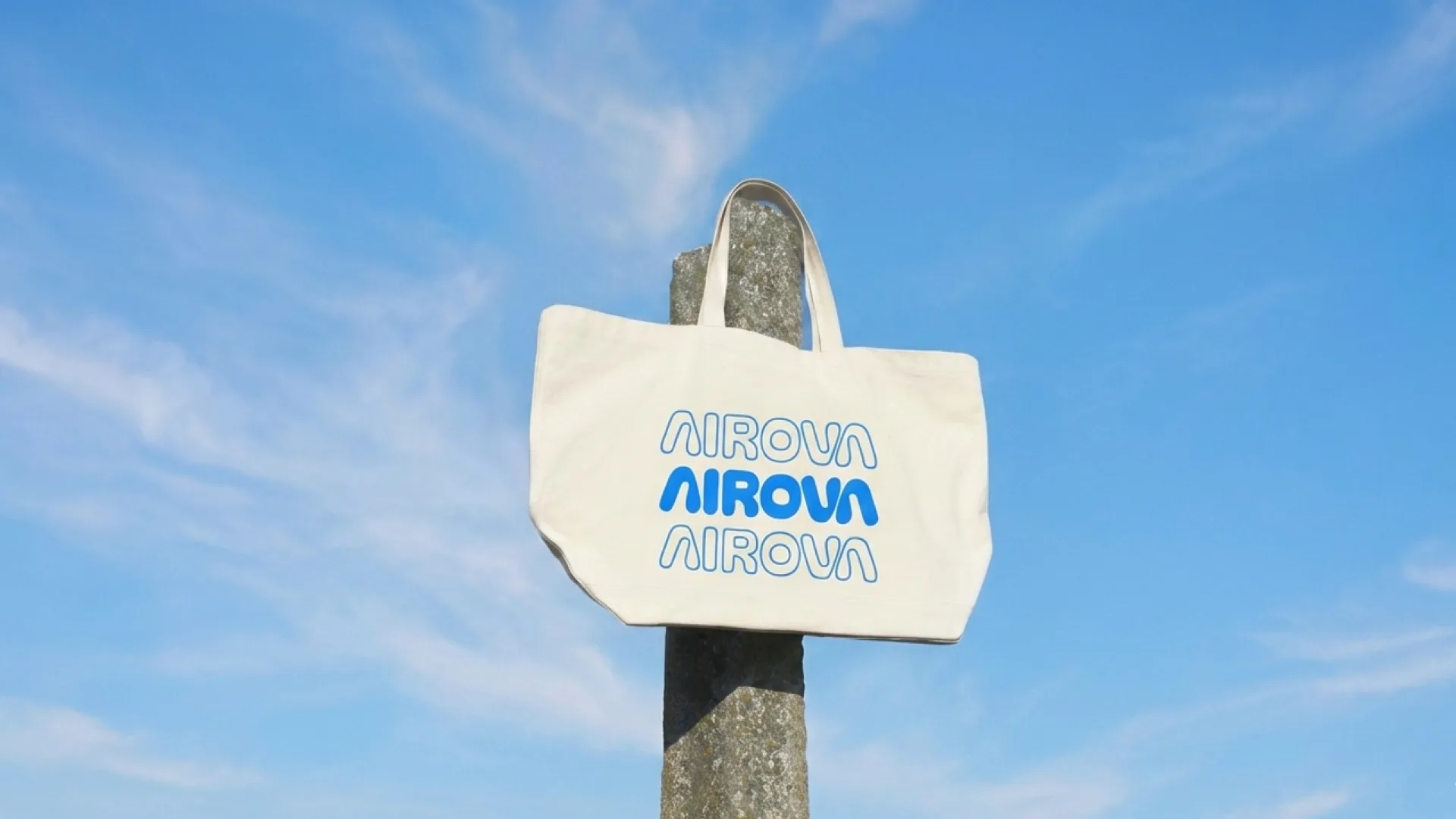 The Airova tote bag, the wordmark repeated three times, hanging from a concrete post against blue sky