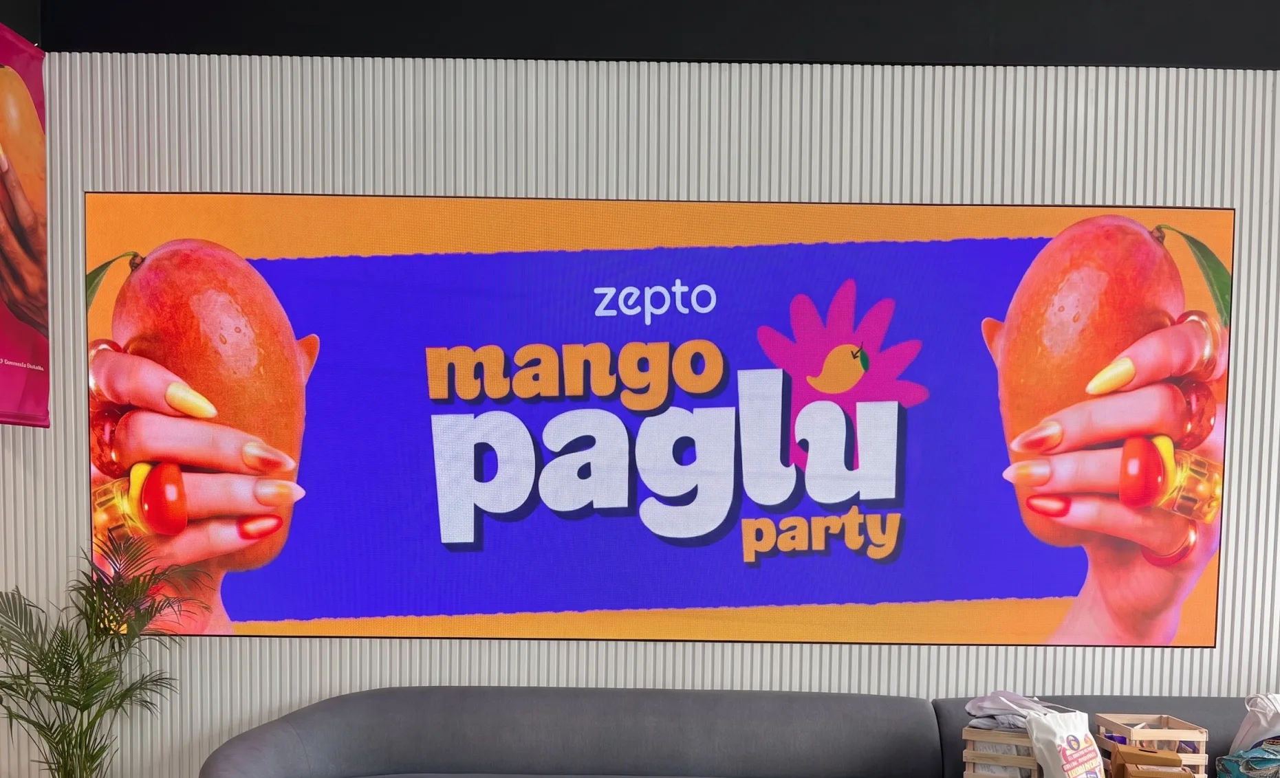 The lockup on the office LED wall, a hand holding a mango either side of it, white and orange type on royal purple inside an orange border