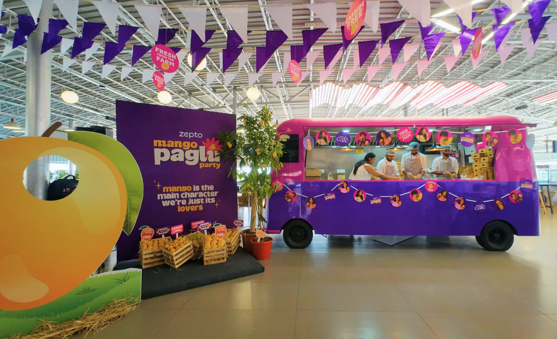 The cafeteria set: a cafe truck repainted pink and purple serving from its window, a giant mango cutout and a purple standee reading mango is the main character, we're just its lovers, crates of mangoes and a mango tree in front, purple and white bunting hung the length of the ceiling