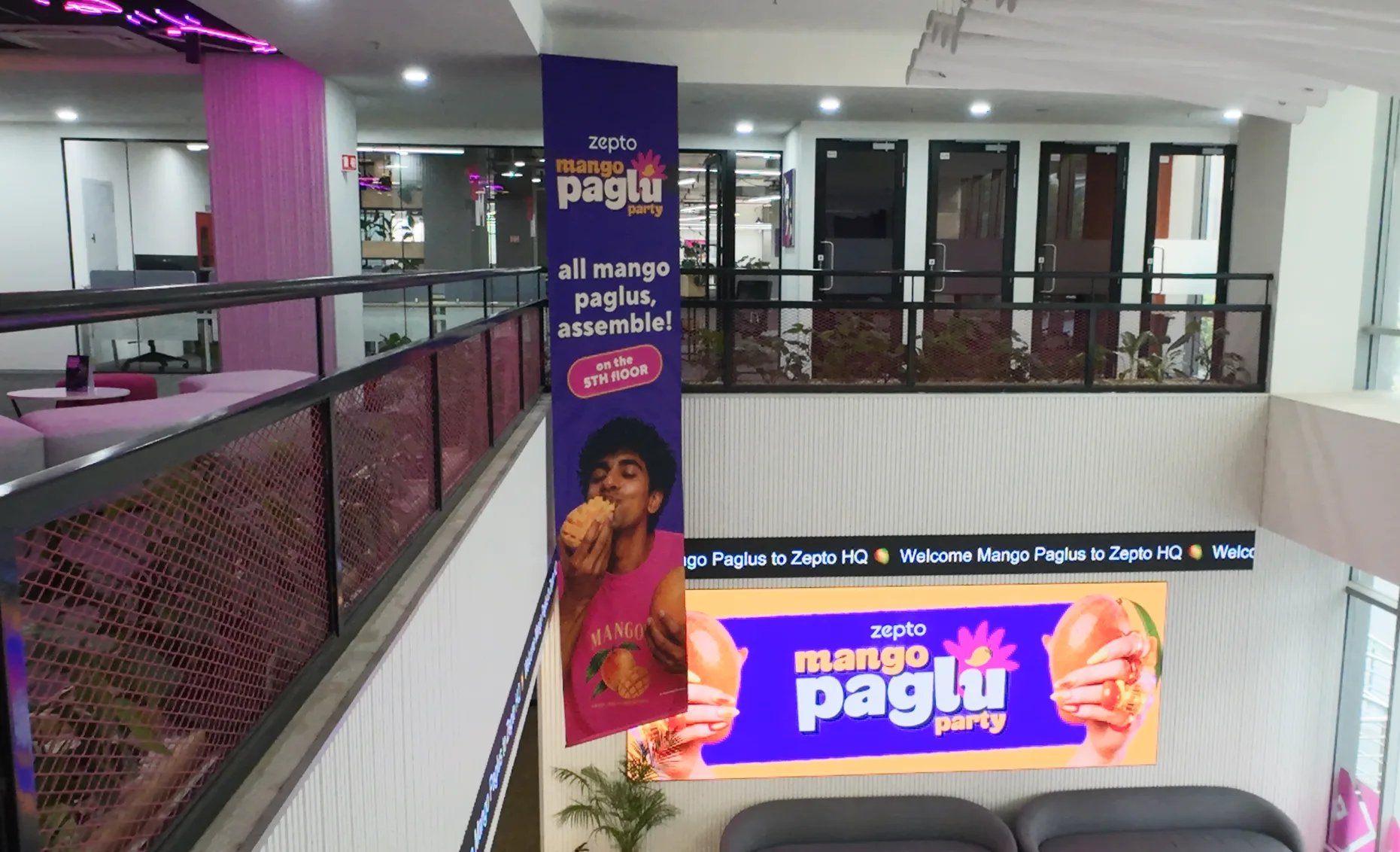 The column banner seen from the mezzanine, the ticker below it reading welcome mango paglus to Zepto HQ, and the lockup running on the screen under that