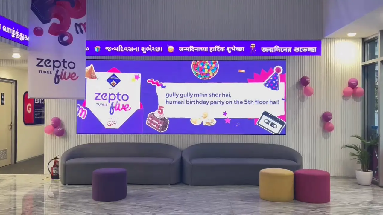 The banner running on the lobby video wall at Zepto HQ, birthday greetings scrolling in Gujarati, Marathi and Bengali along the ticker above it, balloons and a Zepto turns five drop banner either side